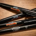 Melnas Watermelon prezentreklāmas pildspalvas ar logo un mājaslapas adreses apdruku uz koka fona.