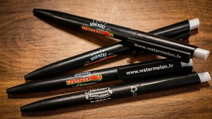 Melnas Watermelon prezentreklāmas pildspalvas ar logo un mājaslapas adreses apdruku uz koka fona.